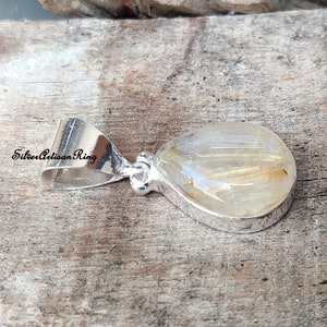 May include: A teardrop-shaped pendant with a clear, golden-hued stone, featuring delicate golden inclusions. The stone is set in a polished silver bezel and bail. The text "Silver Artisan Ring" is visible on the bail.