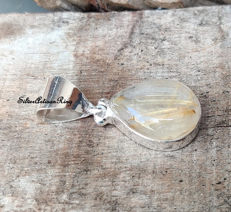 May include: A teardrop-shaped pendant with a clear, golden-hued stone, featuring delicate golden inclusions. The stone is set in a polished silver bezel and bail. The text "Silver Artisan Ring" is visible on the bail.