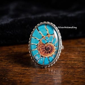 Op de afbeelding: Een ovale ring met een zilveren zetting met geschulpte rand en een centrale cabochon met een spiraalvormige ammoniet fossielafbeelding in tinten helder turkooisblauw en warm bruin. De tekst SilverArtisanRing is rechtsboven zichtbaar.