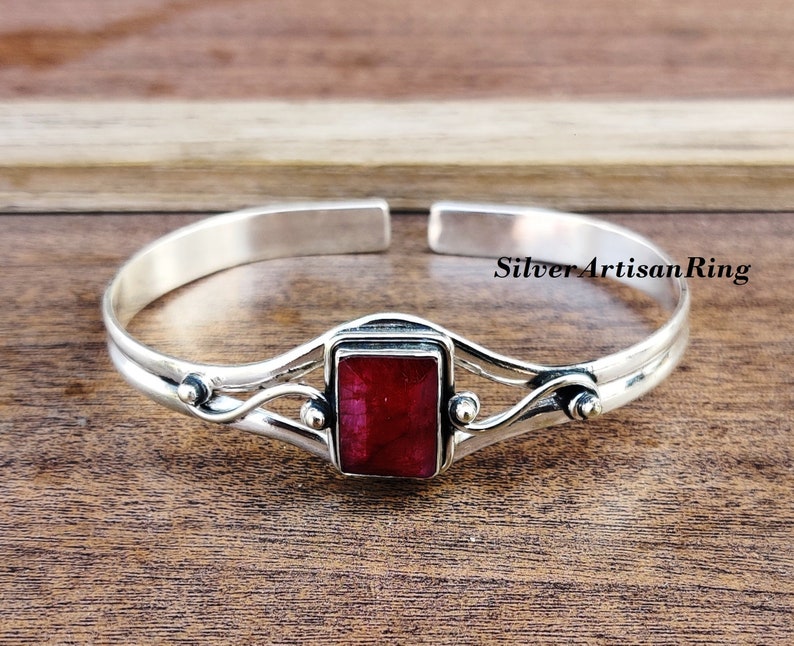 May include: A silver bangle bracelet featuring a red gemstone set in a silver frame. The bracelet has a delicate, flowing design with small silver beads on either side of the gemstone.