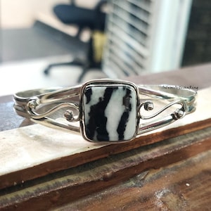 Könnte beinhalten: Ein silberfarbenes Armreif-Armband mit einem rechteckigen, schwarz-weißen Stein. Das Armband hat ein dekoratives Wirbeldesign auf beiden Seiten des Steins. Der Stein hat ein zebraähnliches Muster.