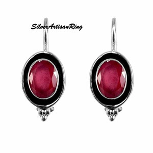 Könnte beinhalten: Ein Paar silberne Ohrringe mit einem roten ovalen Edelstein, der in einer schwarzen Fassung sitzt. Die Ohrringe haben ein schlichtes, elegantes Design.