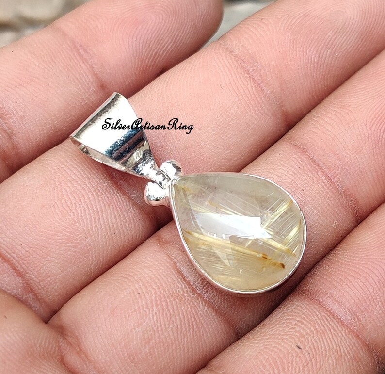 May include: A teardrop-shaped pendant with a silver bail. The pendant features a clear, faceted gemstone with golden inclusions. The gemstone is set in a silver bezel. The text "Silver Artisan Ring" is visible on the image.