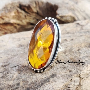 May include: A silver ring with a large, faceted, oval-shaped amber gemstone. The ring has a beaded edge and a smooth, polished band.