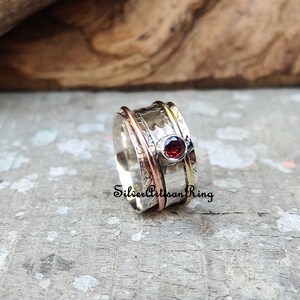 May include: A silver spinning ring with a red gemstone and copper and gold accents. "Silver Artisan Ring" is written on the band.