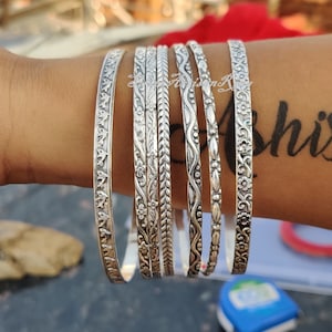 May include: Set of seven silver bangles with intricate floral and geometric designs. The bangles are stacked on a person's wrist.