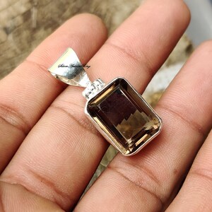Pode incluir: Um pingente de prata com uma pedra preciosa marrom. A pedra preciosa &eacute; cortada em forma retangular com bordas chanfradas. O pingente est&aacute; sobre um fundo branco.