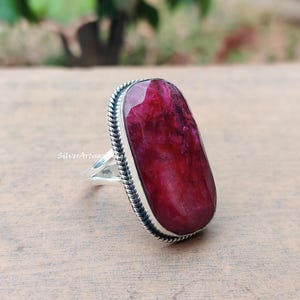 May include: A silver ring with a large, oval-shaped ruby gemstone. The ruby is a rich red hue and is set within a bezel setting with a rope-style border. The ring has a simple, elegant design.