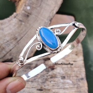 K&ouml;nnte beinhalten: Ein silbernes Armreif-Armband mit einem hellblauen ovalen Edelstein, der in einer silbernen Fassung sitzt. Das Armband hat ein zartes, verziertes Design mit Spiralen und kleinen Perlen.