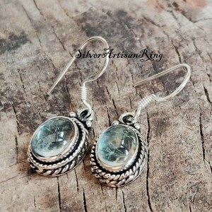 May include: Silver earrings with a light blue oval gemstone set in a silver bezel with a rope-like design. The earrings have a hook closure.