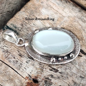 Op de afbeelding: Een zilveren hanger met een witte ovale maansteen, gezet in een getextureerde zilveren bezel. De hanger heeft een lus om aan een ketting te bevestigen.