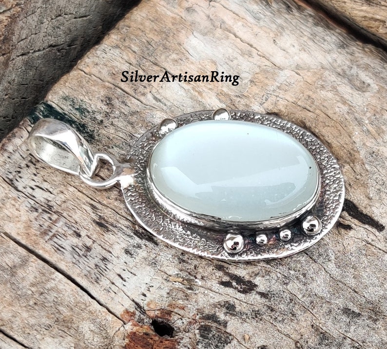 Op de afbeelding: Een zilveren hanger met een witte ovale maansteen, gezet in een getextureerde zilveren bezel. De hanger heeft een lus om aan een ketting te bevestigen.