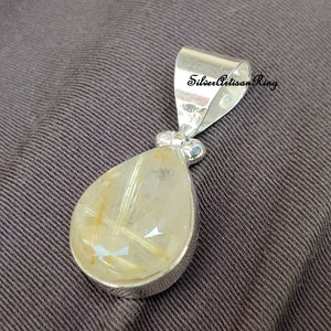 May include: A silver pendant with a teardrop-shaped, translucent gemstone. The gemstone has golden-colored inclusions and is set in a silver bezel. The pendant has a silver bail with the text "Silver Artisan Ring".