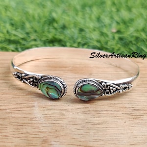 Puede incluir: Un brazalete de plata con dos incrustaciones de concha de abulón en forma de lágrima. El brazalete tiene un delicado diseño floral y está abierto por un lado.