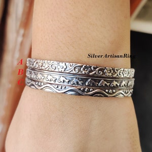 May include: Three silver bangles stacked on a wrist. The top bangle has a floral design, the middle bangle has a dolphin design, and the bottom bangle has a wave design.