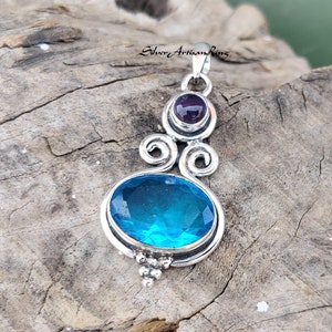 May include: A silver pendant with a blue gemstone and a smaller purple gemstone. The pendant has a swirl design and is set on a wooden background.