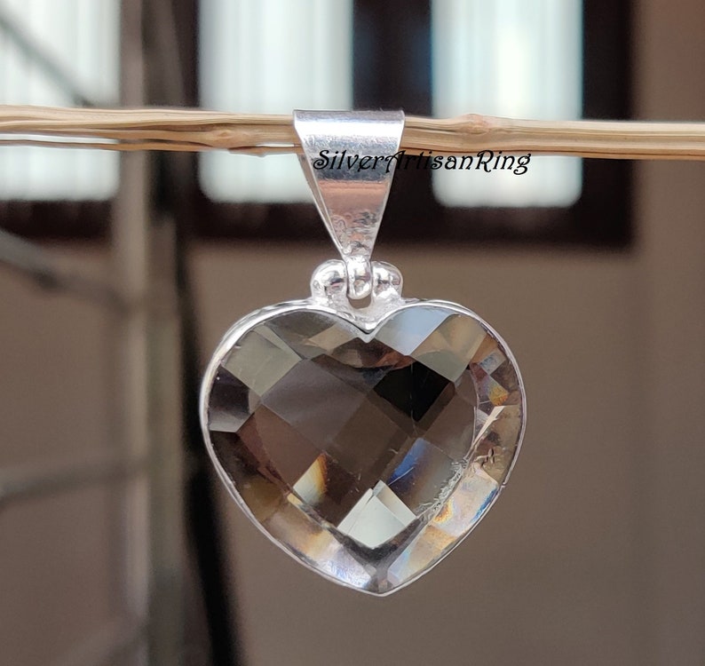 May include: A silver heart-shaped pendant with a clear smoky quartz gemstone. The pendant is hanging from a branch.