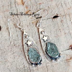 May include: A pair of silver teardrop-shaped earrings with a textured finish. Each earring features a light blue gemstone and a gold star.