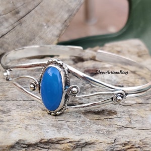 K&ouml;nnte beinhalten: Ein silbernes Armreif-Armband mit einem blauen ovalen Edelstein, der in einem dekorativen silbernen Rahmen gefasst ist. Das Armband hat ein zartes, filigranes Design mit einem Logo von Silver Artisan Ring.