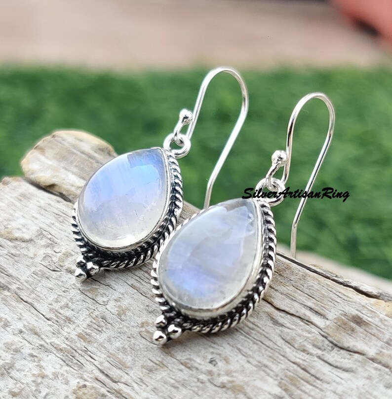 May include: A pair of silver dangle earrings with teardrop-shaped moonstone gemstones. The earrings have a delicate, intricate design with a twisted rope border and small silver beads.