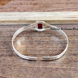 May include: Silver bangle bracelet featuring a red gemstone set in a square bezel. The bracelet is open and has a simple design.