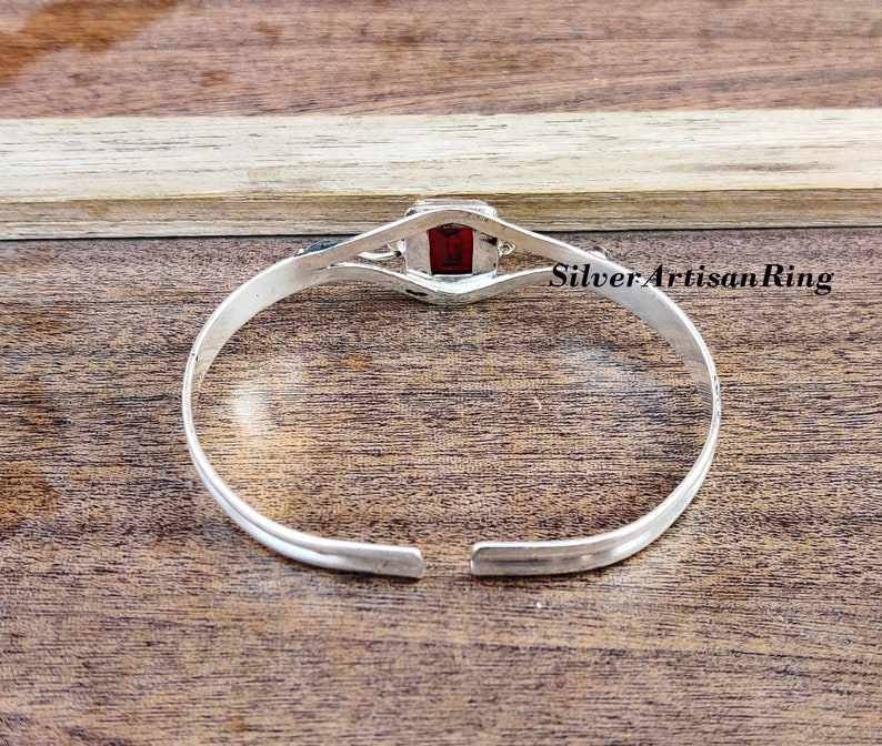 May include: Silver bangle bracelet featuring a red gemstone set in a square bezel. The bracelet is open and has a simple design.