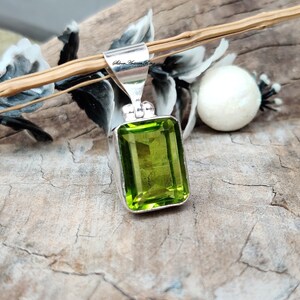 Pode incluir: Pingente retangular de pedra preciosa verde lim&atilde;o com fian&ccedil;a de prata. O pingente &eacute; cravejado em uma moldura de prata e tem uma apar&ecirc;ncia brilhante e facetada. O fundo inclui uma superf&iacute;cie de madeira e elementos decorativos.