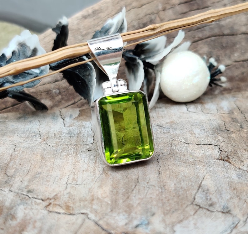 Pode incluir: Pingente retangular de pedra preciosa verde lim&atilde;o com fian&ccedil;a de prata. O pingente &eacute; cravejado em uma moldura de prata e tem uma apar&ecirc;ncia brilhante e facetada. O fundo inclui uma superf&iacute;cie de madeira e elementos decorativos.