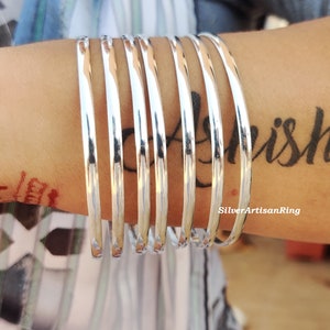 May include: A set of polished silver bangles stacked on a wrist. The bangles are smooth and reflect light. The image also shows a tattoo on the wrist and the text "SilverArtisanRing".