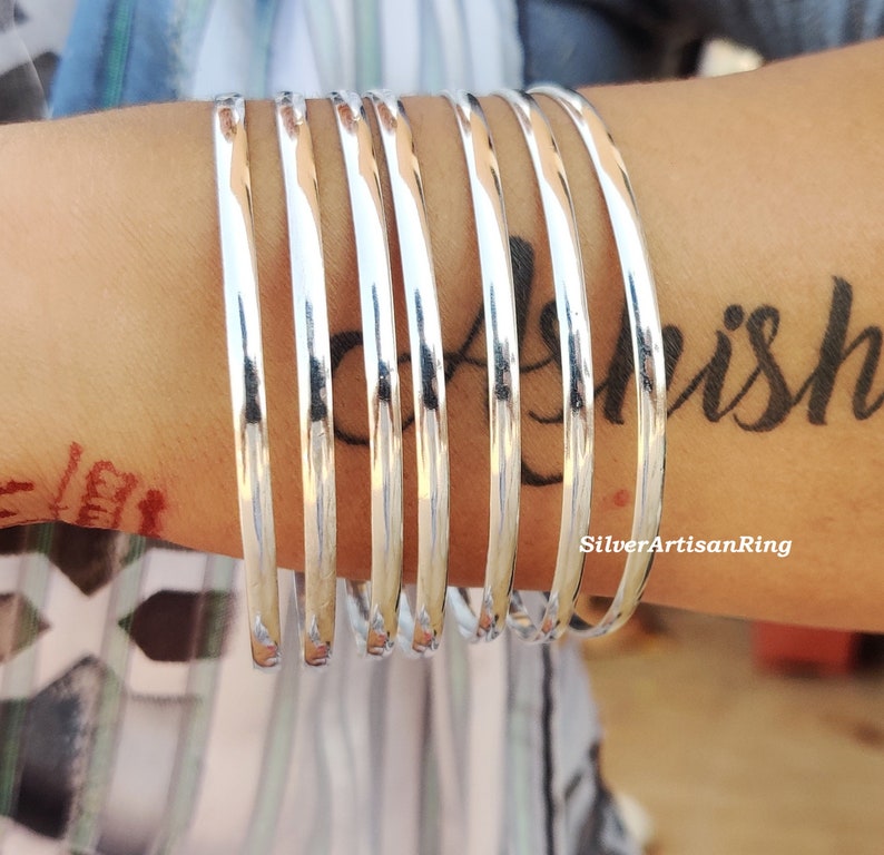 May include: A set of polished silver bangles stacked on a wrist. The bangles are smooth and reflect light. The image also shows a tattoo on the wrist and the text "SilverArtisanRing".