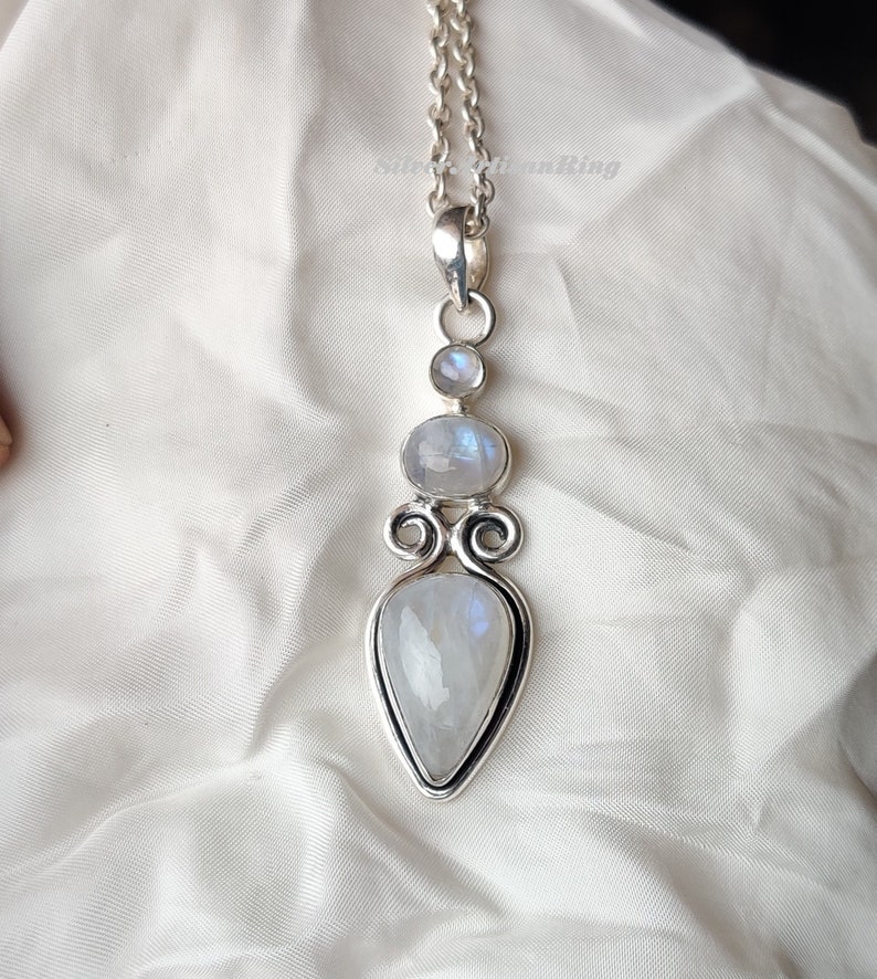 May include: A silver necklace with a teardrop-shaped moonstone pendant. The pendant is surrounded by two smaller moonstones and intricate silver detailing.
