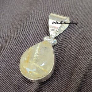 May include: A silver pendant with a teardrop-shaped, translucent gemstone. The gemstone has golden-colored inclusions and is set in a silver bezel. The pendant has a silver bail with the text "Silver Artisan Ring".