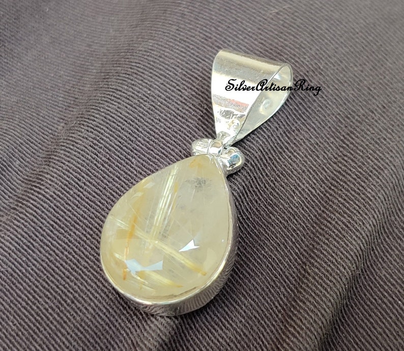 May include: A silver pendant with a teardrop-shaped, translucent gemstone. The gemstone has golden-colored inclusions and is set in a silver bezel. The pendant has a silver bail with the text "Silver Artisan Ring".