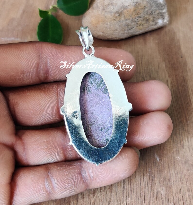 May include: Silver pendant with a purple and green stone set in an oval bezel. The pendant is on a hand, and the back of the pendant is visible. The pendant has a silver bail.