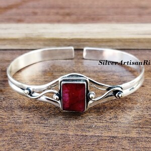 May include: A silver bangle bracelet featuring a red gemstone set in a silver frame. The bracelet has a delicate, flowing design with small silver beads on either side of the gemstone.