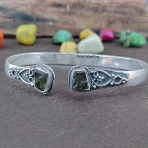 Könnte beinhalten: Ein silberfarbenes Armreif-Armband mit aufwendigen Details und zwei grünen Edelstein-Akzenten. Das offene Armband wird auf einer dunklen Oberfläche mit bunten Steinen im Hintergrund präsentiert. Maße in Zentimetern.