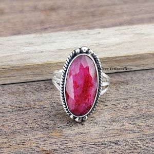 Könnte beinhalten: Ein silberner Ring mit einem großen, ovalen, facettierten Rubin-Edelstein. Der Ring hat ein gedrehtes Band und einen perlenbesetzten Rand um den Stein.