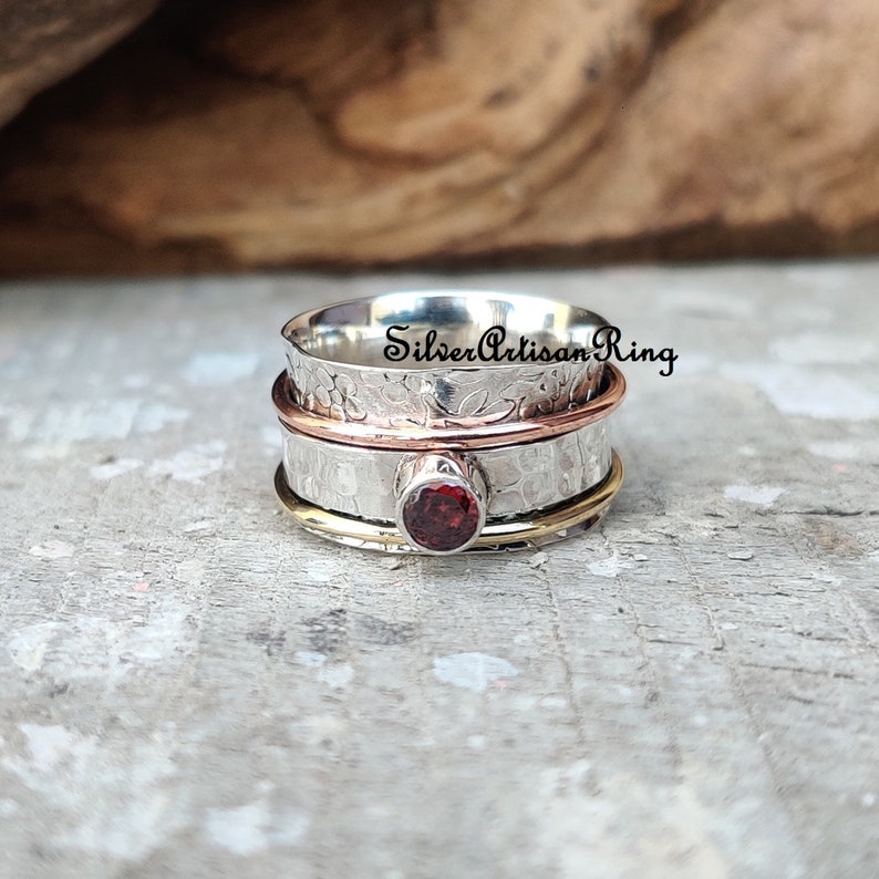 May include: A silver, copper, and gold spinning ring with a red gemstone. The ring is textured and has a floral design.