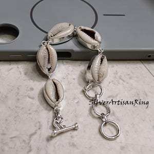 Può includere: Un bracciale in argento con conchiglie di ciprea ovali. Ogni conchiglia è incastonata in una lunetta d'argento e collegata da maglie d'argento. Il bracciale ha una chiusura a levetta. Il testo "SilverArtisanRing" è visibile.