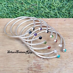 Pode incluir: Um conjunto de oito pulseiras de prata com pedras preciosas de diferentes cores. As pulseiras est&atilde;o empilhadas umas sobre as outras, com as pedras preciosas viradas para cima. As pedras preciosas s&atilde;o das seguintes cores: preto, roxo, verde, vermelho, branco e turquesa.