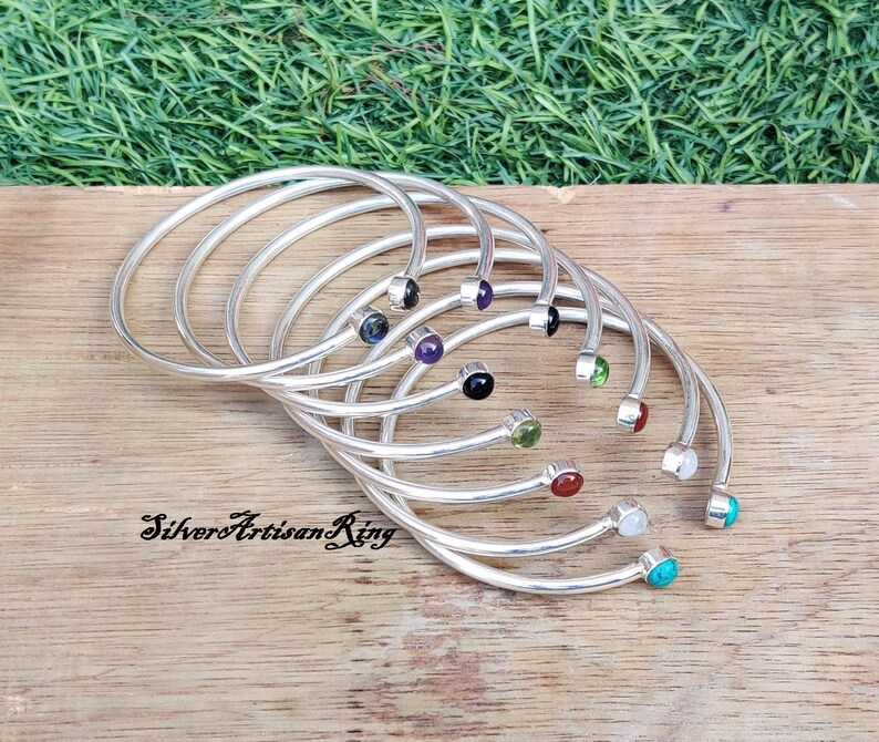 Pode incluir: Um conjunto de oito pulseiras de prata com pedras preciosas de diferentes cores. As pulseiras est&atilde;o empilhadas umas sobre as outras, com as pedras preciosas viradas para cima. As pedras preciosas s&atilde;o das seguintes cores: preto, roxo, verde, vermelho, branco e turquesa.