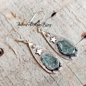 May include: Silver teardrop earrings with a rough aquamarine stone and a gold star accent.  The earrings have a textured finish and are made with sterling silver.