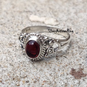 May include: A silver ring with an oval, deep red gemstone. The ring has an ornate design with decorative elements around the stone and on the band. The text "SilverArtisanRing" is visible on the image.
