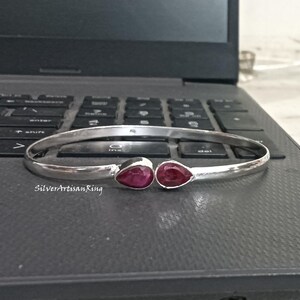 Könnte beinhalten: Ein silberfarbenes Armband mit zwei tropfenförmigen, tiefroten Edelsteinen. Das offene Armband liegt auf einer dunkelgrauen Laptop-Tastatur. Der Text "SilverArtisanRing" ist darunter zu sehen.