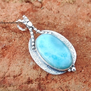 May include: A silver pendant featuring a light blue oval-shaped gemstone with a swirling pattern. The pendant is set in a silver frame with a textured design.