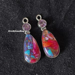 Könnte beinhalten: Ein Paar silberne Ohrhänger mit einem tropfenförmigen Stein. Der Stein ist eine Mischung aus roten, orangen, blauen und violetten Farben. Die Oberseite jedes Ohrrings ist mit einem kleinen violetten Edelstein verziert.
