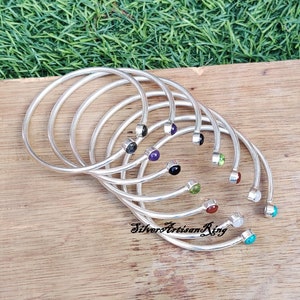 Pode incluir: Um conjunto de sete pulseiras de prata com pedras preciosas de diferentes cores. As pedras preciosas s&atilde;o engastadas no centro de cada pulseira. As pulseiras est&atilde;o empilhadas umas sobre as outras.
