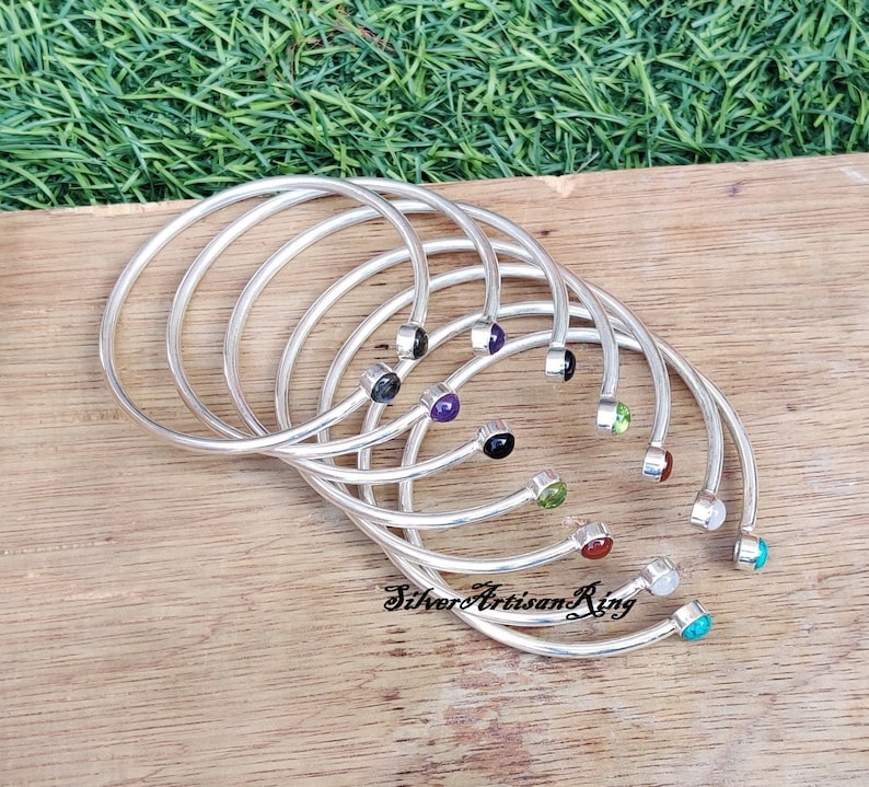 Pode incluir: Um conjunto de sete pulseiras de prata com pedras preciosas de diferentes cores. As pedras preciosas s&atilde;o engastadas no centro de cada pulseira. As pulseiras est&atilde;o empilhadas umas sobre as outras.