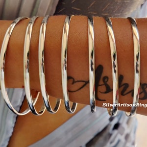 May include: A set of polished silver bangles. The smooth, circular bracelets are shown on an arm, reflecting light. The bangles have a simple, elegant design, suitable for daily wear. The image also shows a tattoo on the arm. The bracelets are from SilverArtisanRing.