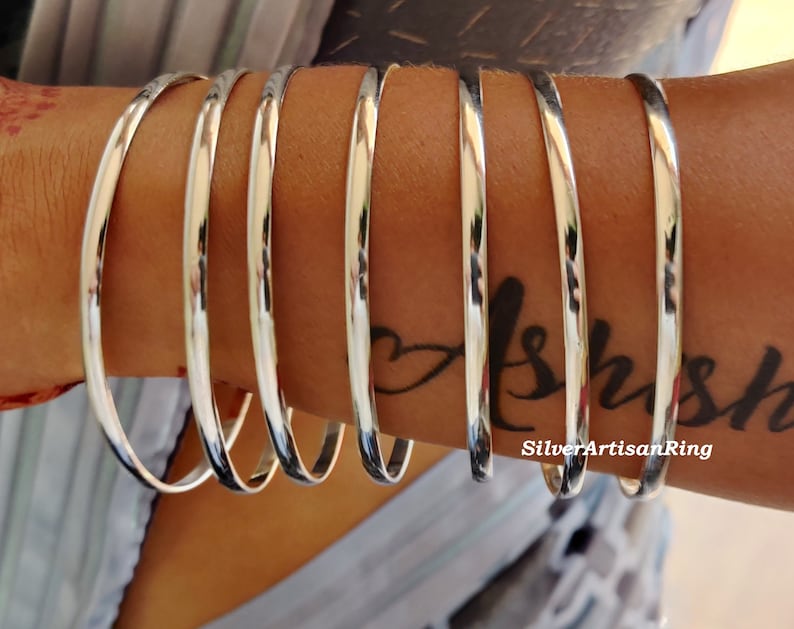 May include: A set of polished silver bangles. The smooth, circular bracelets are shown on an arm, reflecting light. The bangles have a simple, elegant design, suitable for daily wear. The image also shows a tattoo on the arm. The bracelets are from SilverArtisanRing.
